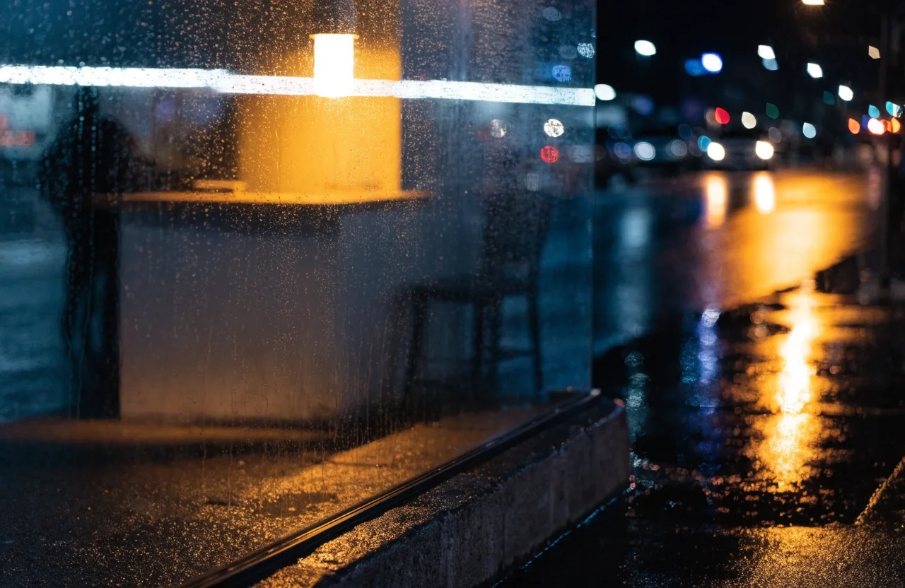 Ventana de un local en San José con lluvia y una lámpara cálida encendida adentro